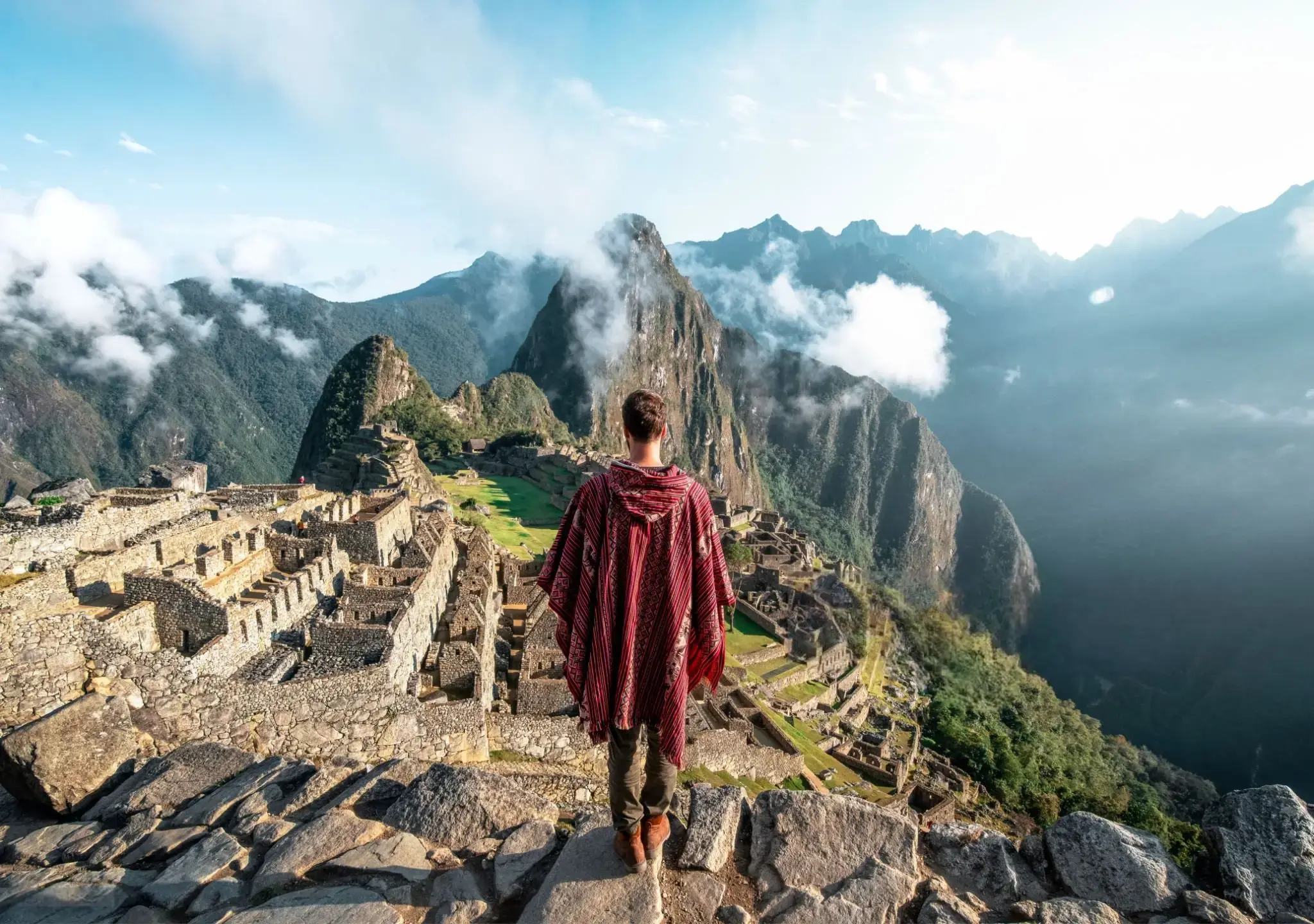 Turista con poncho andino contempla la ciudadela de Machu Picchu, con el Huayna Picchu al fondo entre nubes.